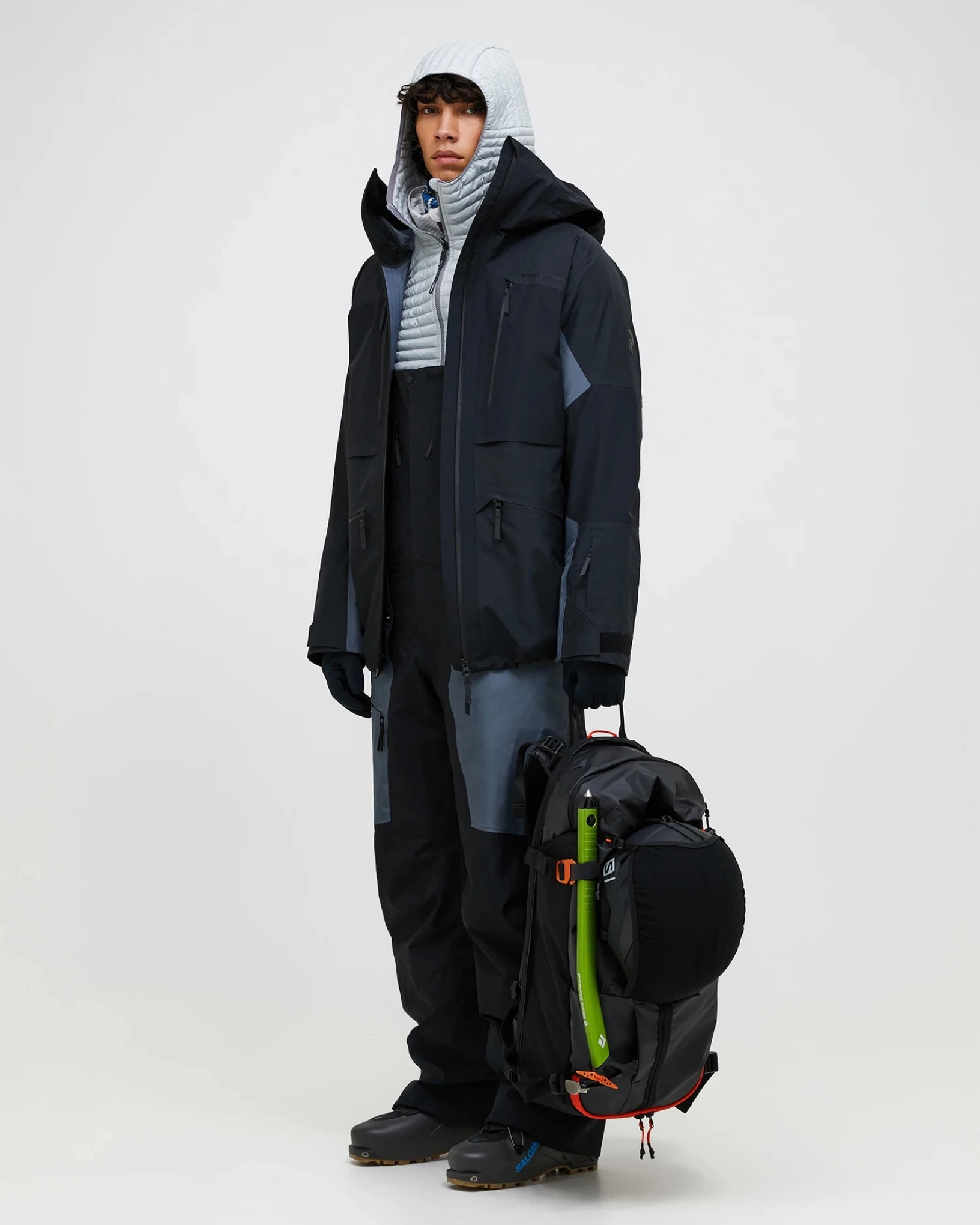 Skier in a dark Ski Supply shell jacket and bib pants, holding a backpack