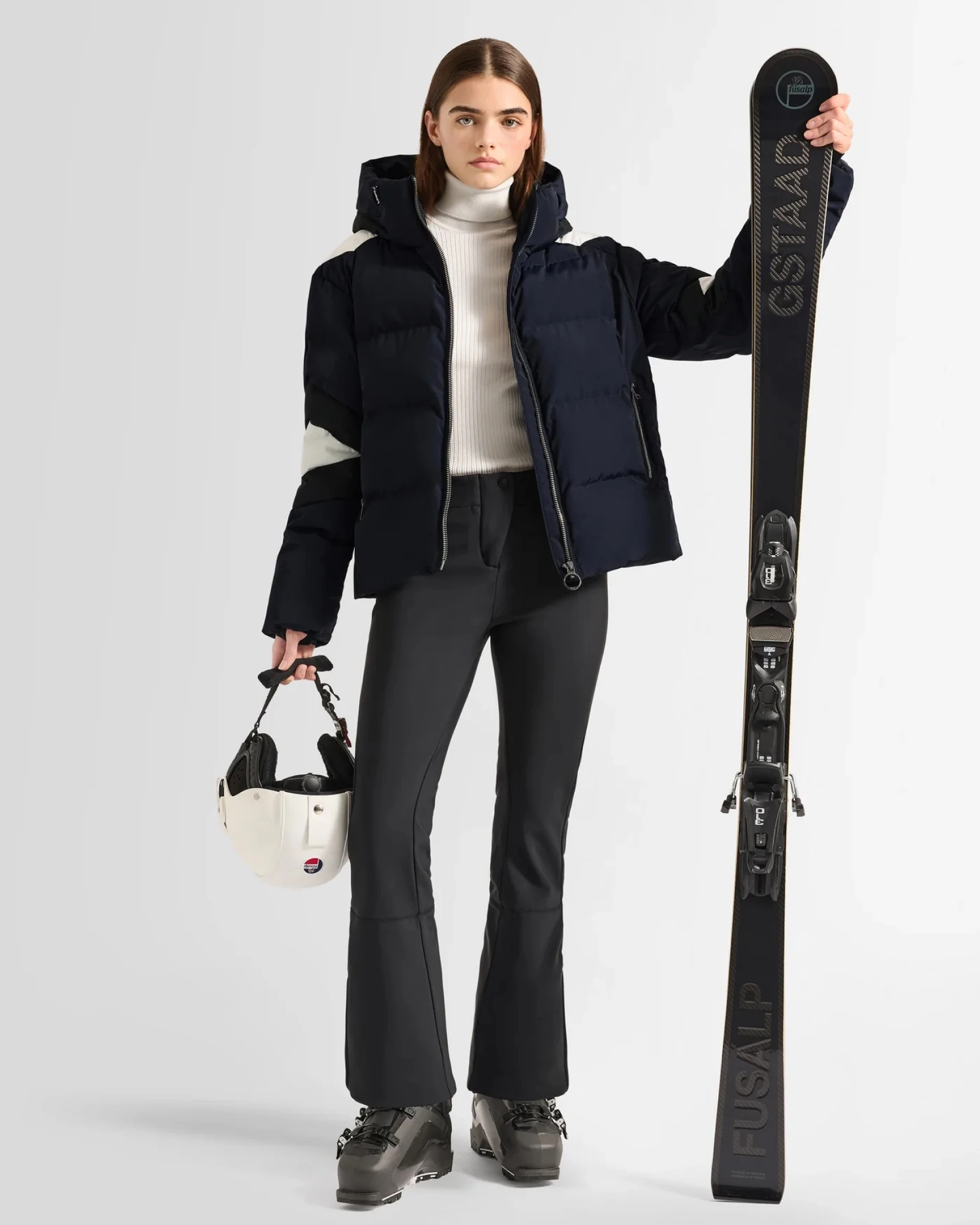 Skier in a navy down jacket, holding a helmet and a pair of skis